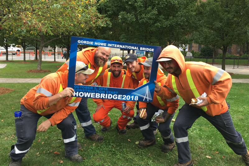 Six workers standing behind a blue Gordie Howe International Bridge frame.